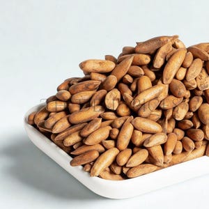 Puede incluir: Un cuenco rectangular blanco rebosante de piñones. Las nueces son de color marrón claro y tienen forma alargada. El fondo es de color azul claro liso.