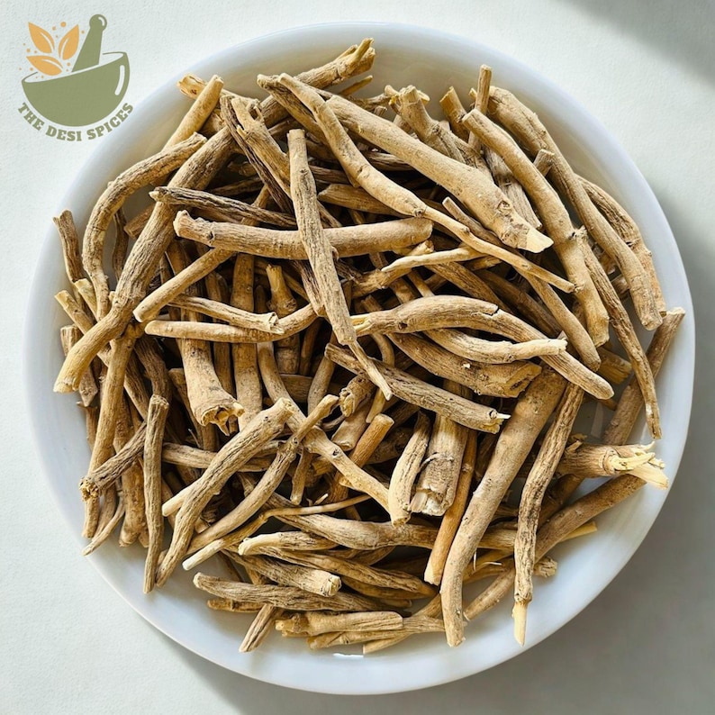 May include: A white bowl filled with dried, beige-colored ashwagandha roots. The roots are long, thin, and have a rough texture. The bowl is set against a light background, with the text "The Desi Spices" and a mortar and pestle logo in the upper left corner.
