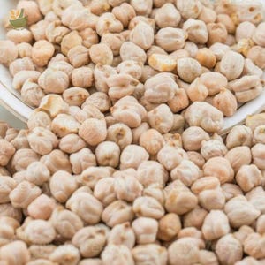 May include: A close-up shot of a pile of dried chickpeas. The chickpeas are beige and tan in color, with a slightly rough texture. They are scattered across the frame, suggesting a bulk quantity. The image is well-lit, highlighting the natural color and shape of the legumes.