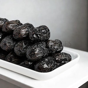 May include: A collection of dark brown dates, with wrinkled skin, presented in a white rectangular dish. The dates have a glossy appearance. The dish is placed on a white surface, with a light grey backdrop.