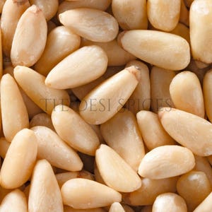 Könnte beinhalten: Nahaufnahme von geschälten Pinienkernen, die eine Ansammlung von cremeweißen, länglichen Samen zeigen. Der Text "THE DESI SPICES" ist sichtbar. Diese Nüsse sind eine gängige Zutat in verschiedenen Küchen.