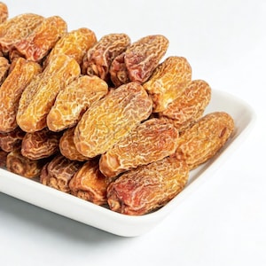 May include: A white rectangular dish filled with a pile of dried dates. The dates are a mix of golden brown and reddish-brown hues, with a wrinkled texture. The dates are arranged in a slightly overflowing heap.