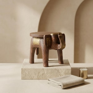 Puede incluir: Un taburete de madera marrón con forma de elefante y una superficie superior plana y circular. El taburete presenta intrincados patrones tallados en las patas y el cuerpo, con detalles en color dorado en la cabeza, orejas y paneles laterales.
