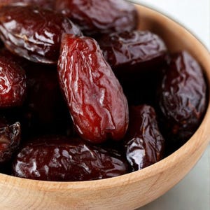 May include: A wooden bowl filled with dark brown dates. The dates have a wrinkled texture and a glossy sheen, suggesting they are fresh and ripe. The bowl is made of light-coloured wood, providing a contrast to the dark fruit.