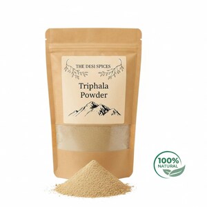 Puede incluir: Una bolsa de papel marrón de Triphala en polvo con una ventana transparente, un pequeño montón de polvo delante. La etiqueta dice "The Desi Spices" y "Triphala Powder" con una ilustración de montaña. A la derecha hay una pegatina verde y blanca "100% Natural".