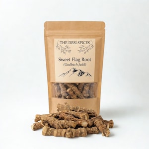 May include: A brown paper bag labeled "THE DESI SPICES" containing Sweet Flag Root (Gudbach Jadd). The bag is partially transparent, revealing the dried root pieces. A pile of the same root pieces is in front of the bag.