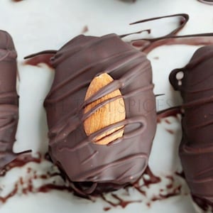May include: Close-up of chocolate-covered treats with an almond on top. The dark chocolate is drizzled over the treats, creating a visually appealing pattern. The treats are arranged on a white surface, suggesting a dessert or snack.