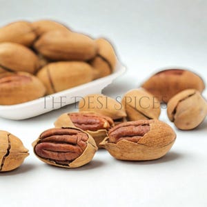 Peut inclure: Gros plan sur des noix de pécan décortiquées et non décortiquées. Les noix de pécan sont de couleur brun clair, certaines étant encore dans leur coquille. Le texte "THE DESI SPICES" est visible en arrière-plan. Les noix de pécan sont disposées sur une surface blanche.