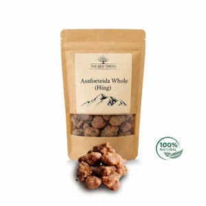 May include: A brown paper bag with a clear window displays asafoetida whole (hing) spices. The label reads "The Desi Spices" and "100% Natural". A small pile of the spice sits in front of the bag.