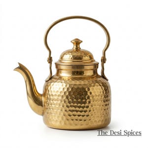 Può includere: Una teiera color oro con una texture martellata. La teiera ha un beccuccio curvo, un manico e un coperchio con un piccolo pomello. Il testo "The Desi Spices" è in basso a destra.