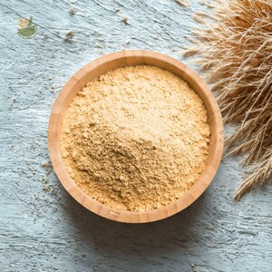 May include: A wooden bowl filled with a light brown powder, possibly a spice or supplement, sits on a weathered, light blue wooden surface. Dried, tan-colored grasses are arranged to the right of the bowl, adding a natural element to the composition.