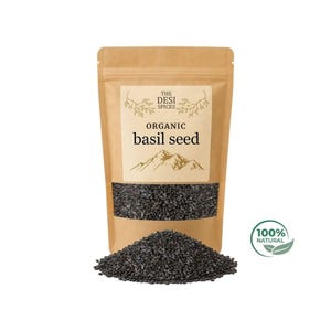 Op de afbeelding: Een bruine papieren zak met biologische basilicumzaden met een doorzichtig venster, met de zwarte zaden. De zak heeft een label met de tekst "The Desi Spices" en "Organic basil seed". Een hoop zaden ligt voor de zak. Een groene en witte "100% Natural" sticker staat rechts.