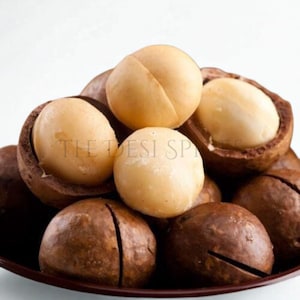 Può includere: Primo piano di noci di macadamia, alcune sgusciate e altre nei loro gusci marroni. Le noci sono di colore beige crema e i gusci sono marrone scuro. Il testo "THE DESI SP" è visibile sullo sfondo.