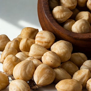 Puede incluir: Primer plano de un cuenco de madera rebosante de avellanas peladas. Las nueces son de color marrón claro con una superficie ligeramente texturizada. Algunas nueces están esparcidas alrededor del cuenco sobre una superficie blanca. La imagen destaca el color y la textura naturales de las nueces.