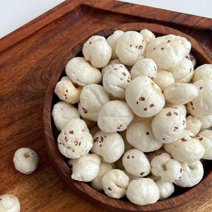 Könnte beinhalten: Eine Holzschale gefüllt mit weißen, gepufften Lotus-Samen, auch bekannt als Fuchsnüsse, mit kleinen braunen Flecken. Die Schale steht auf einem Holztablett. Die Samen sind ein beliebter Snack.