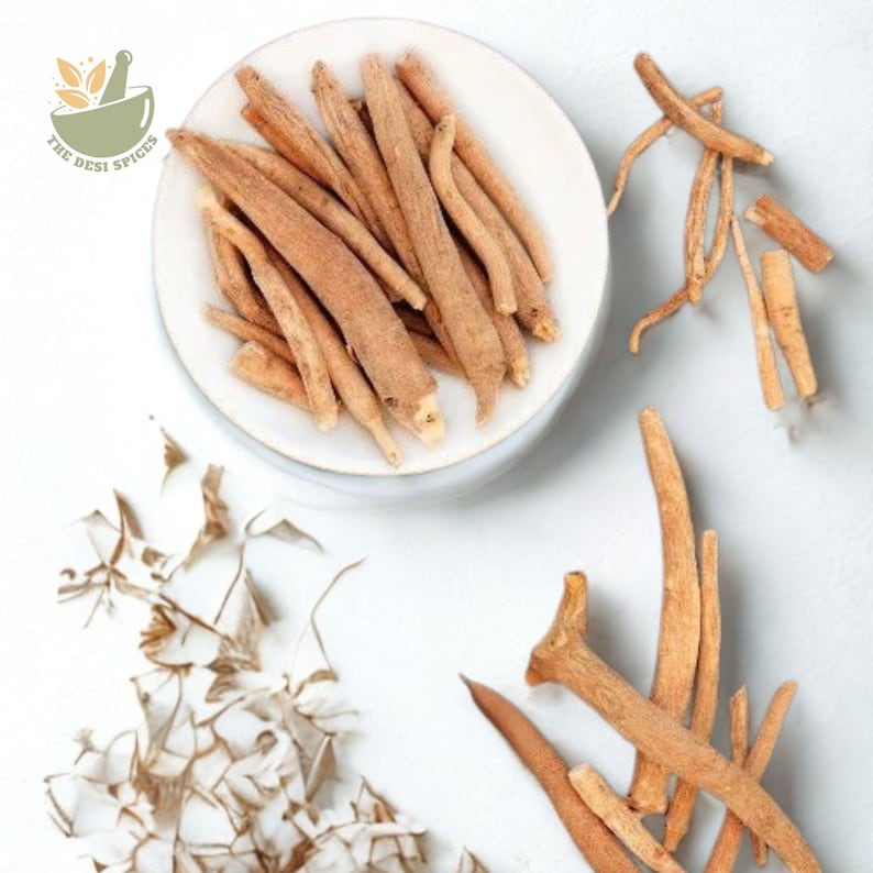 May include: A top-down shot of ashwagandha roots, some on a white plate and others scattered on a white surface. The roots are a light brown color and have a slightly rough texture. The Desi Spices logo is in the upper left corner.