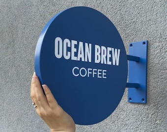 May include: A circular blue metal sign with the white text OCEAN BREW COFFEE printed in the center. The sign is attached to a rectangular blue mounting bracket with two horizontal bars extending from the side of the circular face.