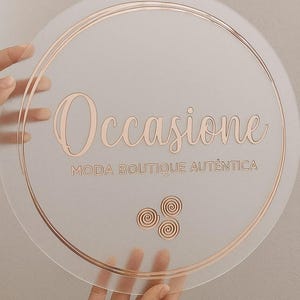 K&ouml;nnte beinhalten: Ein klarer, runder Aufsteller mit ros&eacute;goldfarbenen Buchstaben und Akzenten. Auf dem Schild steht "Occasione" &uuml;ber "Moda Boutique Autentica". Drei Spiralmotive befinden sich darunter. Das Schild wird von H&auml;nden gehalten.