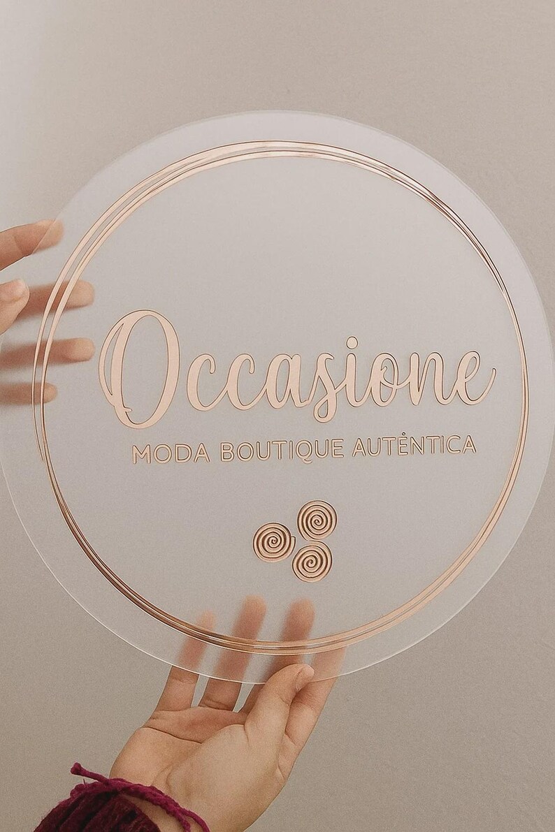 K&ouml;nnte beinhalten: Ein klarer, runder Aufsteller mit ros&eacute;goldfarbenen Buchstaben und Akzenten. Auf dem Schild steht "Occasione" &uuml;ber "Moda Boutique Autentica". Drei Spiralmotive befinden sich darunter. Das Schild wird von H&auml;nden gehalten.