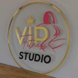 K&ouml;nnte beinhalten: Rundes Schild mit goldenem Rand und der Aufschrift "VIP Fitness STUDIO". Der Text ist in Gold, Pink und Schwarz gehalten. Auf der rechten Seite befindet sich eine pinkfarbene Silhouette einer Person beim Training. Oben befindet sich eine goldene Krone.