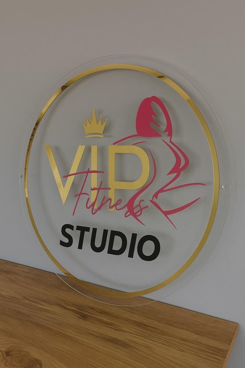 K&ouml;nnte beinhalten: Rundes Schild mit goldenem Rand und der Aufschrift "VIP Fitness STUDIO". Der Text ist in Gold, Pink und Schwarz gehalten. Auf der rechten Seite befindet sich eine pinkfarbene Silhouette einer Person beim Training. Oben befindet sich eine goldene Krone.