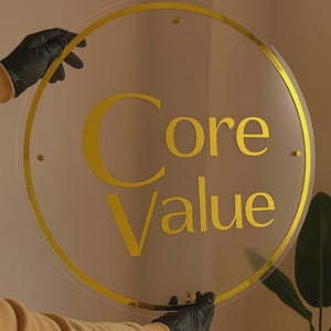 K&ouml;nnte beinhalten: Ein klares, kreisf&ouml;rmiges Schild mit goldenem Rand und dem Schriftzug "Core Value" in goldener Schrift. Das Schild wird von behandschuhten H&auml;nden gehalten. Der neutrale Hintergrund deutet auf ein Gesch&auml;fts- oder Branding-Thema hin.