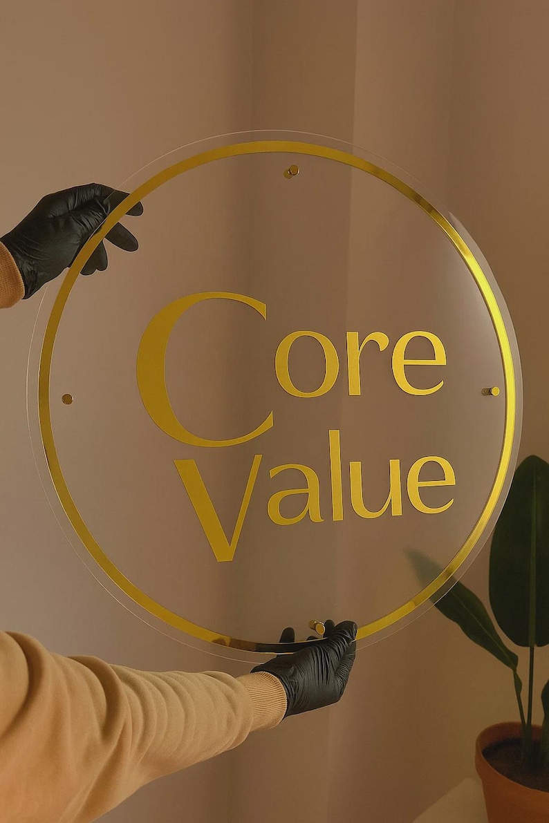 K&ouml;nnte beinhalten: Ein klares, kreisf&ouml;rmiges Schild mit goldenem Rand und dem Schriftzug "Core Value" in goldener Schrift. Das Schild wird von behandschuhten H&auml;nden gehalten. Der neutrale Hintergrund deutet auf ein Gesch&auml;fts- oder Branding-Thema hin.