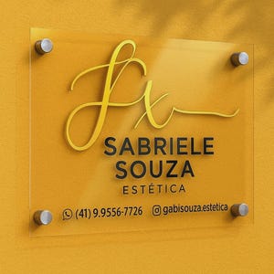 K&ouml;nnte beinhalten: Klarer Acrylschild mit goldfarbenen Buchstaben und silbernen Befestigungen. Das Schild zeigt "Sabriele Souza Est&eacute;tica" mit Telefonnummer und Instagram-Handle. Der Hintergrund ist einfarbig gelb.