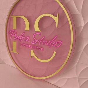 K&ouml;nnte beinhalten: Ein rundes, rosa Schild mit goldenem Rand und Schriftzug. Das Schild zeigt "DC Pudra Studio" in einer kursiven Schriftart, mit den Worten "студия краси" darunter. Der Hintergrund ist eine strukturierte, hellrosa Wand.
