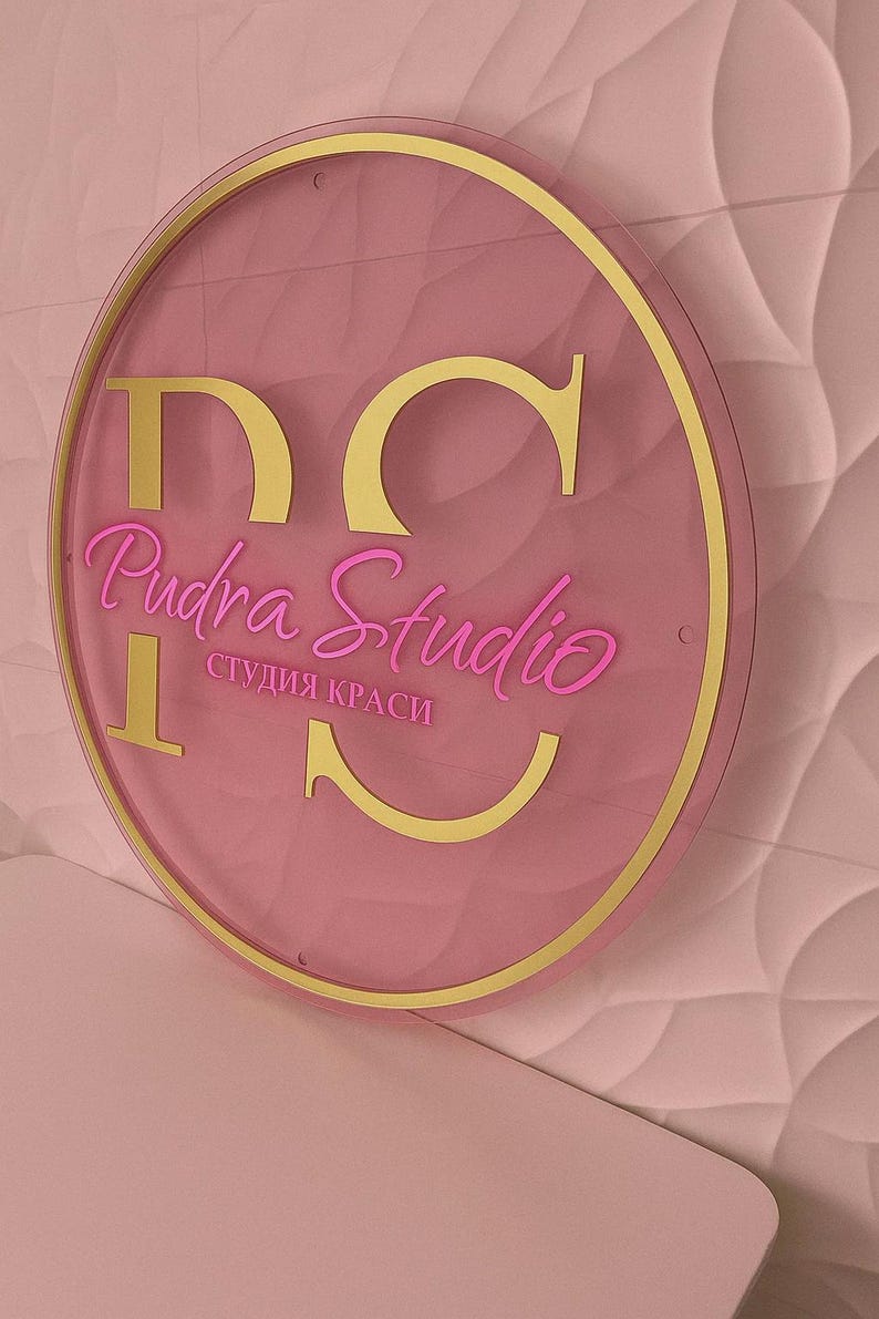 K&ouml;nnte beinhalten: Ein rundes, rosa Schild mit goldenem Rand und Schriftzug. Das Schild zeigt "DC Pudra Studio" in einer kursiven Schriftart, mit den Worten "студия краси" darunter. Der Hintergrund ist eine strukturierte, hellrosa Wand.