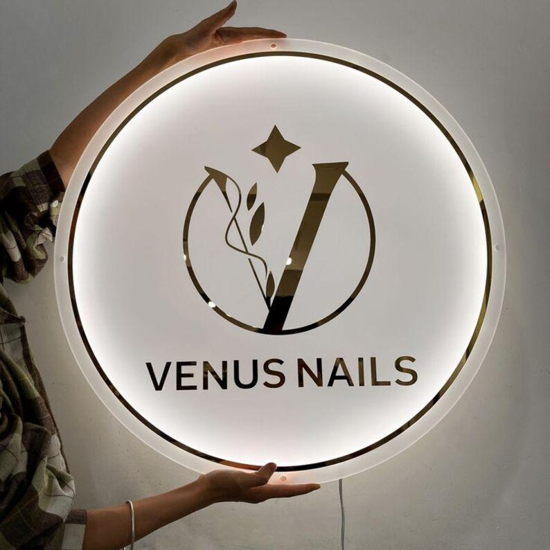 K&ouml;nnte beinhalten: Ein rundes, beleuchtetes Schild mit dem Text "VENUS NAILS". Das Schild hat einen goldfarbenen Rand und ein zentrales Design mit einem Stern, einer "V"-Form und floralen Elementen. Das Schild wird von einer Person gehalten.