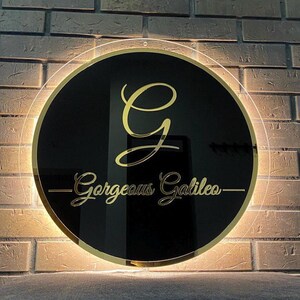 K&ouml;nnte beinhalten: Ein runder, schwarzer Leuchtschild mit goldenem Rand, einem goldenen Buchstaben "G" und dem Schriftzug "Gorgeous Galileo" in Gold. Das Schild wird von einem warmwei&szlig;en Licht beleuchtet und an einer Backsteinmauer befestigt.