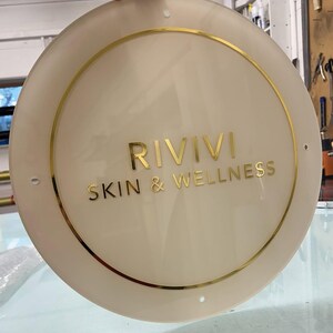 K&ouml;nnte beinhalten: Ein cremefarbenes rundes Schild mit goldfarbenen Buchstaben und einem goldenen Rand. Auf dem Schild steht "RIVIVI SKIN & WELLNESS". Das Schild wird von einer Hand gehalten.