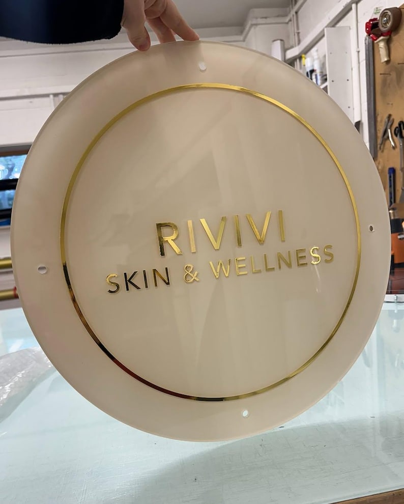 K&ouml;nnte beinhalten: Ein cremefarbenes rundes Schild mit goldfarbenen Buchstaben und einem goldenen Rand. Auf dem Schild steht "RIVIVI SKIN & WELLNESS". Das Schild wird von einer Hand gehalten.