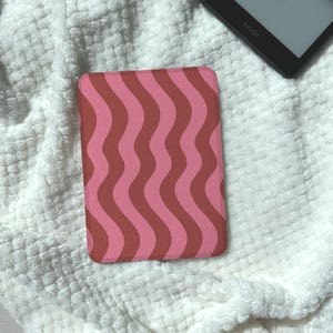 Könnte beinhalten: Eine Kindle-Hülle mit einem wellenförmigen Muster in Rosa- und Rottönen. Die rechteckige Hülle ist für einen E-Reader konzipiert. Die Hülle liegt auf einer weißen, strukturierten Oberfläche, im Hintergrund ist ein Kindle-E-Reader zu sehen.