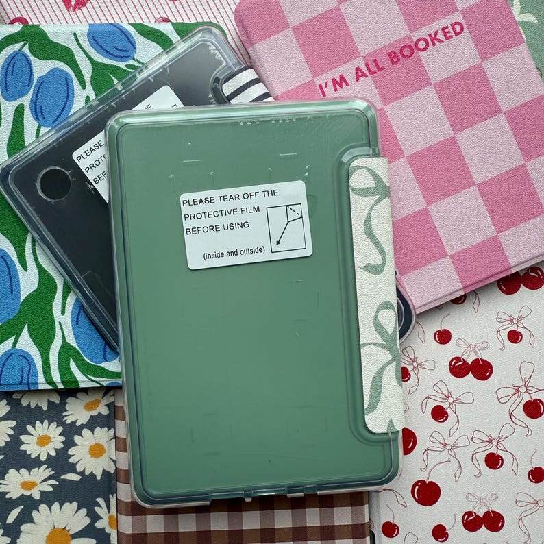 Puede incluir: Una funda verde para lector electr&oacute;nico con una pegatina de pel&iacute;cula protectora, rodeada de varias cubiertas de libros. La pegatina dice "PLEASE TEAR OFF THE PROTECTIVE FILM BEFORE USING." Otras cubiertas presentan patrones florales, a cuadros y de cerezas. El texto "I'M ALL BOOKED" es visible.