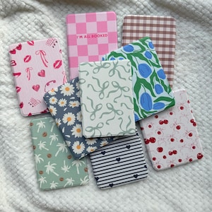 May include: A selection of patterned notebooks with diverse designs. The notebooks display patterns like pink gingham, daisies, cherries, bows, and stripes. One notebook has the text "I'M ALL BOOKED". The notebooks are arranged on a white, textured surface.