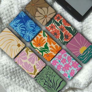Könnte beinhalten: Eine Sammlung von zehn rechteckigen E-Reader-Hüllen mit verschiedenen farbenfrohen Mustern, darunter blaue Monstera-Blätter, orangefarbene und rosa Blumen, eine gelbe Sonne über Wellen und abstrakte Blumendesigns auf einer weißen, strukturierten Oberfläche.