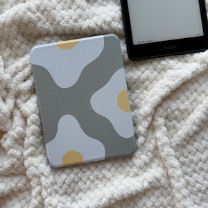 Może przedstawiać: Prostokątne etui na Kindle z szarym tłem i wzorem białych i żółtych abstrakcyjnych kształtów. Etui leży obok czarnego urządzenia Kindle na białej, teksturowanej powierzchni.