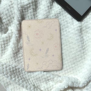 May include: A light pink Kindle case with a celestial design featuring suns, moons, stars, clouds, and hands holding feathers. The case is rectangular and has a soft, textured appearance. A Kindle device is partially visible in the background.