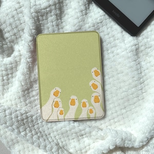 Puede incluir: Una funda Kindle verde claro con un diseño de gansos blancos con picos amarillos. La funda rectangular tiene un acabado liso. Un dispositivo Kindle es visible en el fondo.