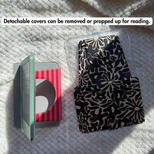 May include: A light green e-reader with a red and pink striped stand, alongside a detachable black and beige floral patterned cover. The text "Detachable covers can be removed or propped up for reading." is visible at the top of the image.