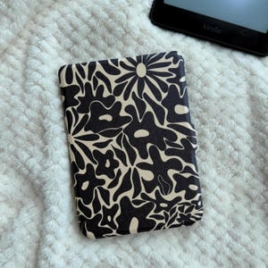 以下が含まれることがあります： 黒とベージュの花柄Kindleケース。ケースは、様式化された花と葉の繰り返しデザインが特徴です。Kindleは背景に部分的に表示されています。