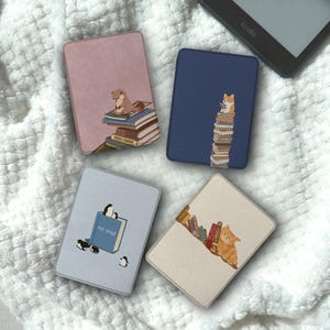 May include: Four rectangular e-reader cases featuring illustrated designs. One pink case shows an otter on books, a navy case features a corgi on a book stack, a light blue case depicts penguins with a book labeled not smut, and a beige case shows a cat.