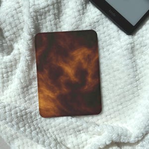 May include: A rectangular Kindle case with a swirling pattern of brown, orange, and yellow hues. The case is resting on a white, textured surface, with a Kindle device visible in the background.