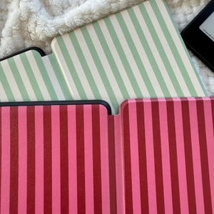 May include: Close-up of three e-reader covers with vertical stripes. One cover is mint green and white, and two are shades of pink and red. The covers are open, revealing the interior. The background is a soft, white, textured fabric.