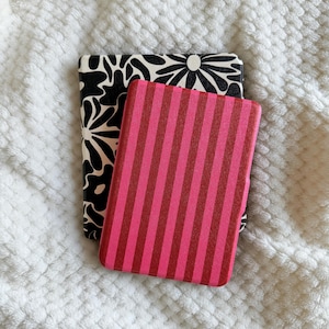 Puede incluir: Dos tarjeteros rectangulares. Uno tiene un estampado floral en blanco y negro. El otro tiene un diseño a rayas rosas y rojas. Ambos descansan sobre una superficie blanca texturizada.