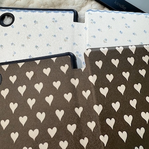 May include: Two e-reader cases with black frames. The top case is white with a small blue floral pattern and a blue scalloped edge. The bottom case is dark brown with a repeating pattern of off-white hearts. Both cases feature a circular cutout.