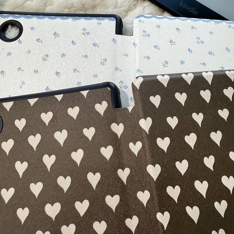 May include: Two e-reader cases with black frames. The top case is white with a small blue floral pattern and a blue scalloped edge. The bottom case is dark brown with a repeating pattern of off-white hearts. Both cases feature a circular cutout.