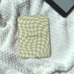 May include: A Kindle cover with a wavy checkerboard pattern in light green and off-white. The cover has the text "in my bookish era" in a script font. The cover is resting on a white, textured blanket, with a Kindle device visible in the background.